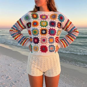 May include: A long-sleeve sweater featuring a white base with a multi-colored granny square pattern in shades of red, yellow, blue, green, and purple, paired with cream-colored crochet shorts with a textured, open-work knit design.