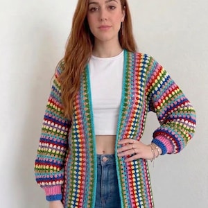 May include: A long-sleeved, open-front crochet cardigan featuring a vibrant, multi-coloured striped pattern in shades of pink, blue, green, yellow, and red. The garment has a solid light blue trim along the front edges and pink ribbed cuffs.