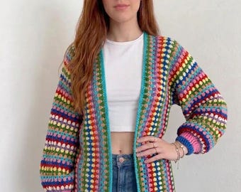 Puede incluir: Un cárdigan de ganchillo de manga larga y frente abierto con un vibrante patrón de rayas multicolor en tonos rosa, azul, verde, amarillo y rojo. La prenda presenta un borde azul claro sólido a lo largo de los bordes frontales y puños acanalados rosas.