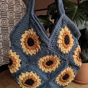 Puede incluir: Una bolsa de mano de crochet con fondo azul y dise&ntilde;os de girasoles. La bolsa tiene dos asas y est&aacute; hecha con un hilo grueso.