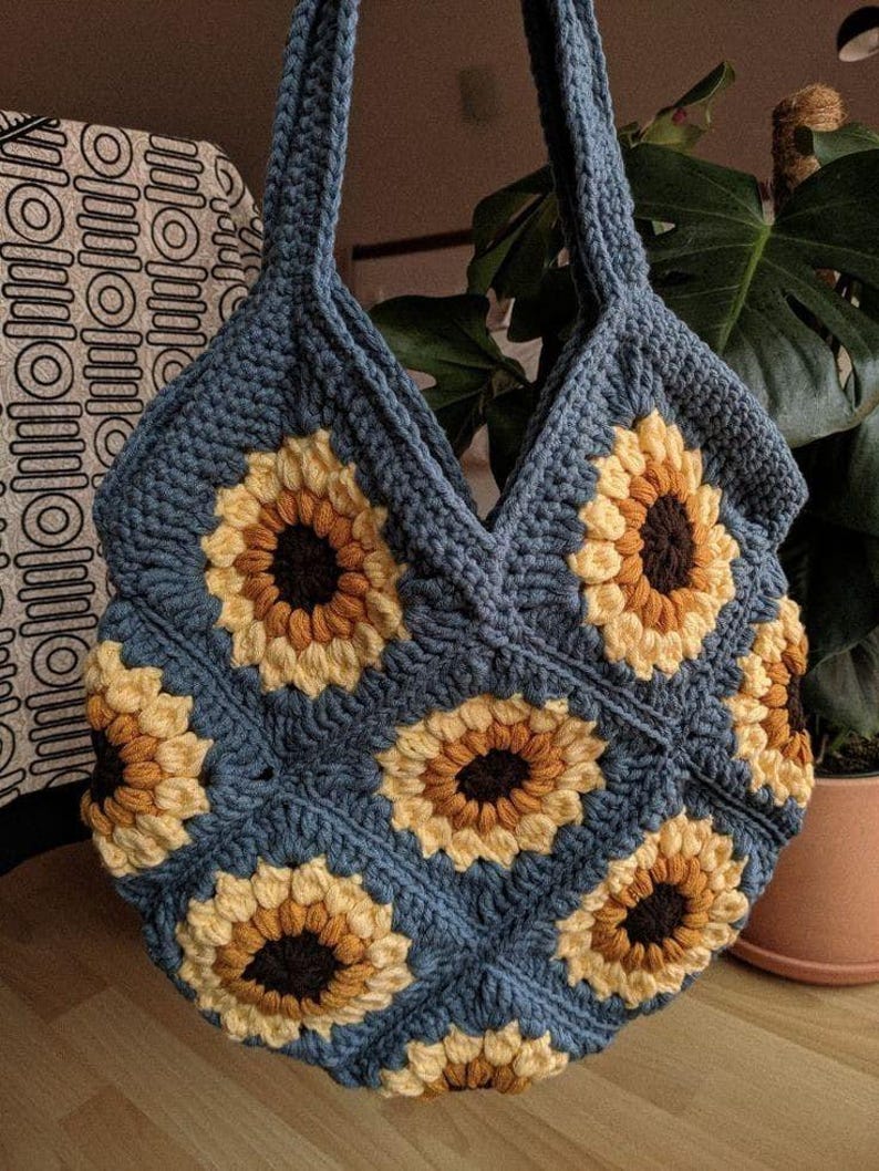 Puede incluir: Una bolsa de mano de crochet con fondo azul y dise&ntilde;os de girasoles. La bolsa tiene dos asas y est&aacute; hecha con un hilo grueso.