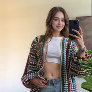 May include: A colorful, long-sleeved crochet cardigan with a square pattern in various colors. The cardigan has a light brown trim and is paired with a white top and light blue jeans. The sleeves are long and puffy.