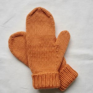 Könnte beinhalten: Ein Paar gestrickte Fäustlinge in einem warmen Orangeton. Die Fäustlinge sind aus dickem Garn gefertigt, mit geripptem Bündchen und abgerundeter Oberseite. Die Fäustlinge liegen auf einer weißen Oberfläche.