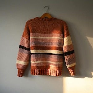 May include: A cozy, striped sweater in shades of orange, pink, purple, and cream hangs on a wooden hanger. The sweater features a textured knit design with horizontal stripes across the body and sleeves. The cuffs and hem are a darker shade of orange.