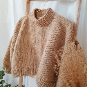 Könnte beinhalten: Ein brauner Strickpullover mit hohem Kragen und lockerer Passform. Der Pullover hängt an einer Holzleiter.