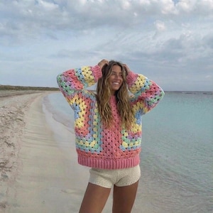 Peut inclure: Un pull crocheté coloré avec un motif de carrés granny. Le pull présente une base rose avec des carrés arc-en-ciel pastel en jaune, bleu et vert. Les manches sont longues et la coupe est décontractée. Le pull est associé à un short crème.