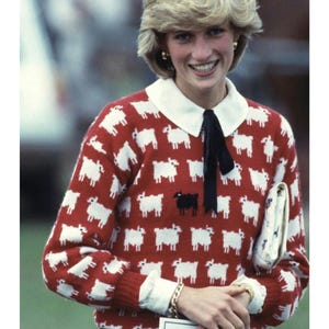May include: Red sweater with white sheep pattern and a single black sheep. The sweater has a white collar and cuffs, and is paired with white pants. A black bow is tied at the collar.