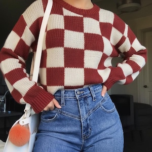 Op de afbeelding: Een rood en crème geblokte trui met lange mouwen. De trui wordt gecombineerd met een jeans met hoge taille. Een witte handtas met een oranje pompon hangt aan de schouder.