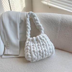 May include: A white, chunky-knit handbag with braided handles. The bag is made of thick, soft yarn and has a rounded shape. A white blazer is partially visible in the background, resting on a textured, off-white surface.