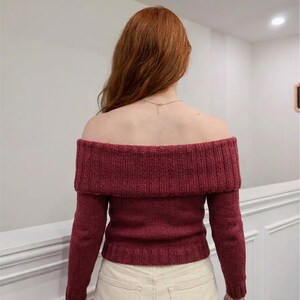 May include: A long-sleeved, off-the-shoulder jumper in a deep maroon colour. The garment features a wide, ribbed fold-over collar that extends across the shoulders and upper back, with matching ribbed cuffs and a ribbed hemline at the waist.