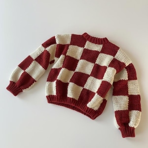 Könnte beinhalten: Ein rot-weiß karierter Strickpullover. Der Pullover hat lange Ärmel mit gerippten Bündchen und Saum. Der Halsausschnitt ist ebenfalls gerippt. Der Pullover ist aus einem weichen Strickmaterial gefertigt.