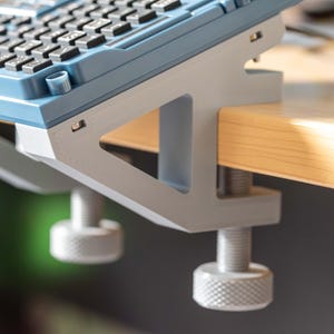 May include: A gray and white keyboard stand with a blue top, clamped to a wooden surface. The stand has two adjustable knobs and a sturdy design, providing support for a keyboard. The stand is designed to elevate a keyboard.