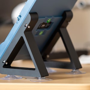 May include: A blue tablet or device is supported by a black stand with a triangular design. The stand has clear suction cups on the bottom for stability. The device has a few buttons and ports visible on the side.