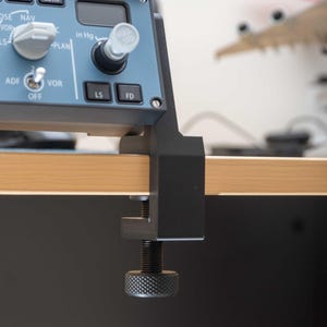 Puede incluir: Primer plano de un panel de instrumentos de avión azul y negro con botones, diales y una pantalla digital. El panel está montado en una superficie de madera con una abrazadera negra. El panel tiene los textos "WPT", "NAV", "VOR", "CSTR", "ADF", "LS" y "FD".