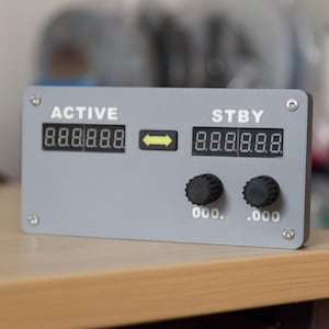 Puede incluir: Un dispositivo electrónico gris con pantallas digitales etiquetadas como "ACTIVE" y "STBY". Cuenta con perillas negras y una flecha amarilla. Las pantallas muestran lecturas numéricas. El dispositivo está montado sobre una superficie de madera.