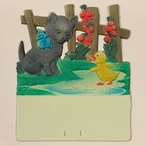 May include: A vintage cardstock notepad with a 3D image of a gray kitten wearing a blue bow and a yellow duckling standing in a pond. The scene is set in front of a brown wooden fence with red flowers. The cardstock is light green and has a blank space for writing. The bottom of the cardstock has the word "Germany" printed on it.