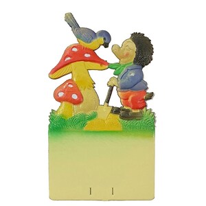May include: A colorful figurine of a hedgehog with a blue jacket and red pants standing next to a red and yellow mushroom. A blue bird is perched on top of the mushroom. The figurine is attached to a yellow and green background with a blank white space for writing.