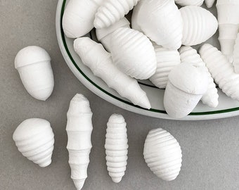 Op de afbeelding: Een verzameling witte, papier-maché eikels en andere natuur-geïnspireerde vormen op een wit bord met een groene rand.