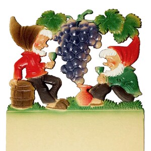 May include: Two gnomes, one wearing red and one wearing green, are standing next to a bunch of purple grapes. The gnome in red is holding a glass of wine and the gnome in green is holding a glass of wine. The gnomes are standing on a green grassy area with a brown barrel and a red pot.