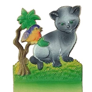 May include: A gray kitten with green eyes sits in a grassy field with a small tree and a blue and orange bird perched on a branch. The scene is on a blank card with two holes at the bottom.