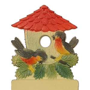 May include: A vintage birdhouse ornament with a red roof, green leaves, and two birds with red breasts. The birdhouse has a blank space for writing or drawing.