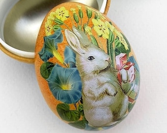May include: A white ceramic Easter egg ornament with a blue ribbon hanger. The egg is decorated with a colorful illustration of a rabbit, flowers, and leaves.