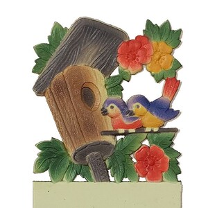 May include: A wooden birdhouse with a brown roof and a bluebird couple perched on a branch. The birdhouse is surrounded by green leaves and red flowers.