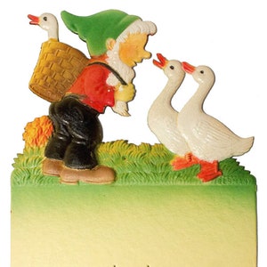 May include: A green-hatted gnome with a basket on his back looks at two white geese with red feet standing on green grass.