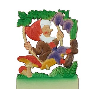 May include: A colorful plastic figurine of a gnome sitting on a mushroom swing. The gnome is wearing a red hat, brown jacket, purple pants, and brown shoes. The swing is made of rope and is attached to a mushroom with green leaves. The figurine is attached to a white card with two holes at the bottom.