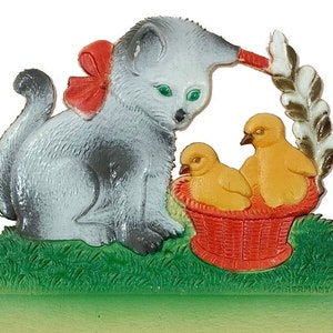 May include: A gray and white kitten with a red bow sits on a green grassy surface, looking at two yellow chicks in a red wicker basket. The basket is decorated with a green leafy branch.