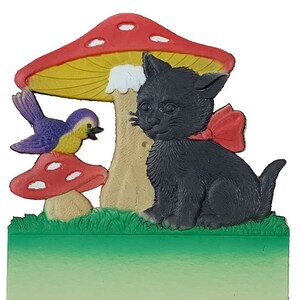 May include: A black cat with a red bow sits under a red and yellow mushroom with white spots. A blue and yellow bird sits on a smaller red mushroom with white spots. The scene is set against a green grassy background.