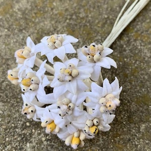 May include: A cluster of white fabric flowers with yellow and tan centers and a bundle of off-white stems. The flowers have a star-like shape and are arranged in a small bouquet. The background is a textured, light gray surface.