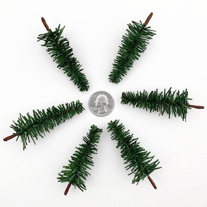 May include: Six miniature green artificial pine trees with brown stems, arranged in a circle around a US quarter.