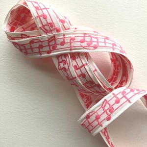 May include: White ribbon with a pink musical note pattern. The ribbon is folded and curved, showcasing the detailed embroidery of the musical notes and staff lines. The ribbon's edges are clean and well-defined, creating a visually appealing decorative element.
