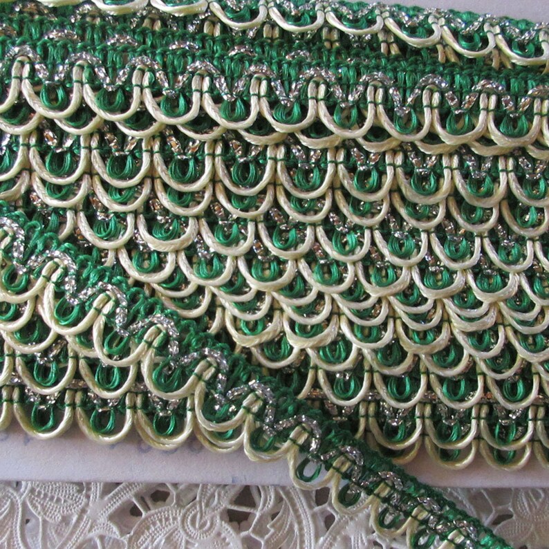 May include: Green and cream trim with silver thread. The trim has a scalloped edge and is made of a woven fabric.