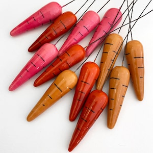 May include: A collection of 12 hand-painted wooden carrots in shades of pink, orange, and brown. The carrots are painted with black lines and are attached to thin wires.