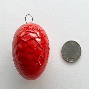 May include: A glossy red, egg-shaped ornament with a textured, scale-like pattern. A small, black wire loop is attached at the top. A U.S. quarter coin is next to the ornament for size comparison.