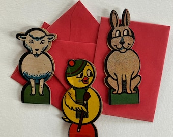 May include: Three vintage-style cardboard cutouts of a lamb, a chick, and a rabbit, each standing on a green base. The lamb has blue accents, the chick wears a hat and scarf, and the rabbit is brown. They are set against a red envelope.