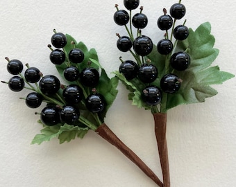 May include: Two artificial black currant berry sprigs with green leaves and brown stems. The berries are glossy and spherical, clustered on thin green stems. The leaves are a muted green, and the stems are wrapped in brown paper.