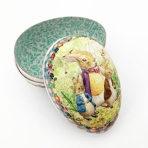 May include: An oval-shaped Easter egg box with a hinged lid. The lid features a colorful illustration of three rabbits in a garden scene, surrounded by a floral border. The interior of the box is patterned with a green floral design.