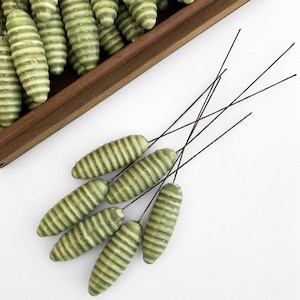 May include: A set of six green, ribbed, artificial flower buds with wire stems. The buds are arranged in a fan shape on a white background.