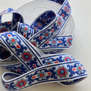 May include: A roll of woven ribbon featuring a repeating floral pattern in shades of blue, light blue, and bright orange on a white background. The decorative trim has a textured surface and is coiled into a circular spool shape for storage.