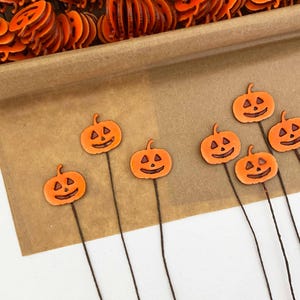 May include: Orange pumpkin-shaped decorations with black facial features on thin brown stems. The Halloween-themed decorations are arranged on a brown surface, with more pumpkins in the background. These are ideal for seasonal crafts and decor.