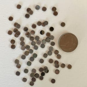 May include: A collection of small, round, metallic discs scattered on a white surface. A U.S. quarter coin is included for size comparison. The discs have a reflective, dark gray appearance, with some showing hints of brown and green.