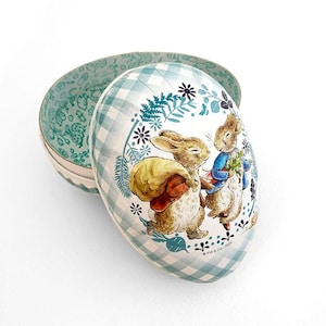 May include: An egg-shaped decorative box with a blue and white checkered pattern. The box features an illustration of two rabbits, one carrying a sack and the other holding vegetables. The interior of the box has a floral design.
