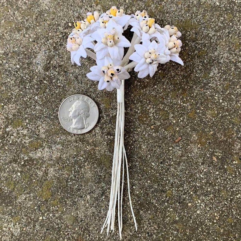 May include: A small bouquet of white paper flowers with yellow and brown centers. The flowers are tied together with white string. A U.S. quarter coin is next to the flowers for size comparison.