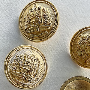 May include: Close-up of several gold-colored buttons. Each button features a detailed embossed design with a coat of arms, laurel leaves, and text. The buttons are round with a ridged edge and a shiny finish.