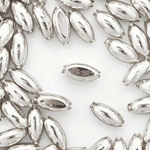 May include: A close-up of a group of silver oval-shaped beads. The beads are scattered on a white surface.