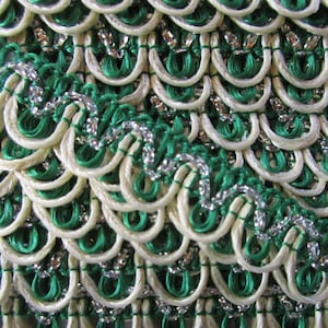 May include: Close-up of a white and green trim with silver accents. The trim is made of woven cord and has a scalloped edge.