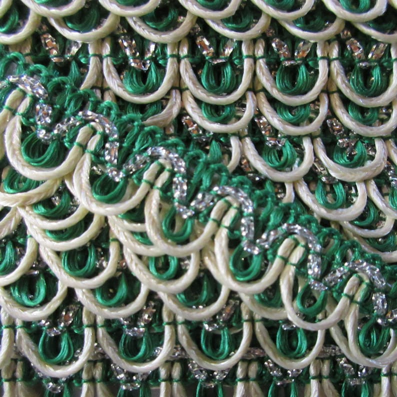 May include: Close-up of a white and green trim with silver accents. The trim is made of woven cord and has a scalloped edge.